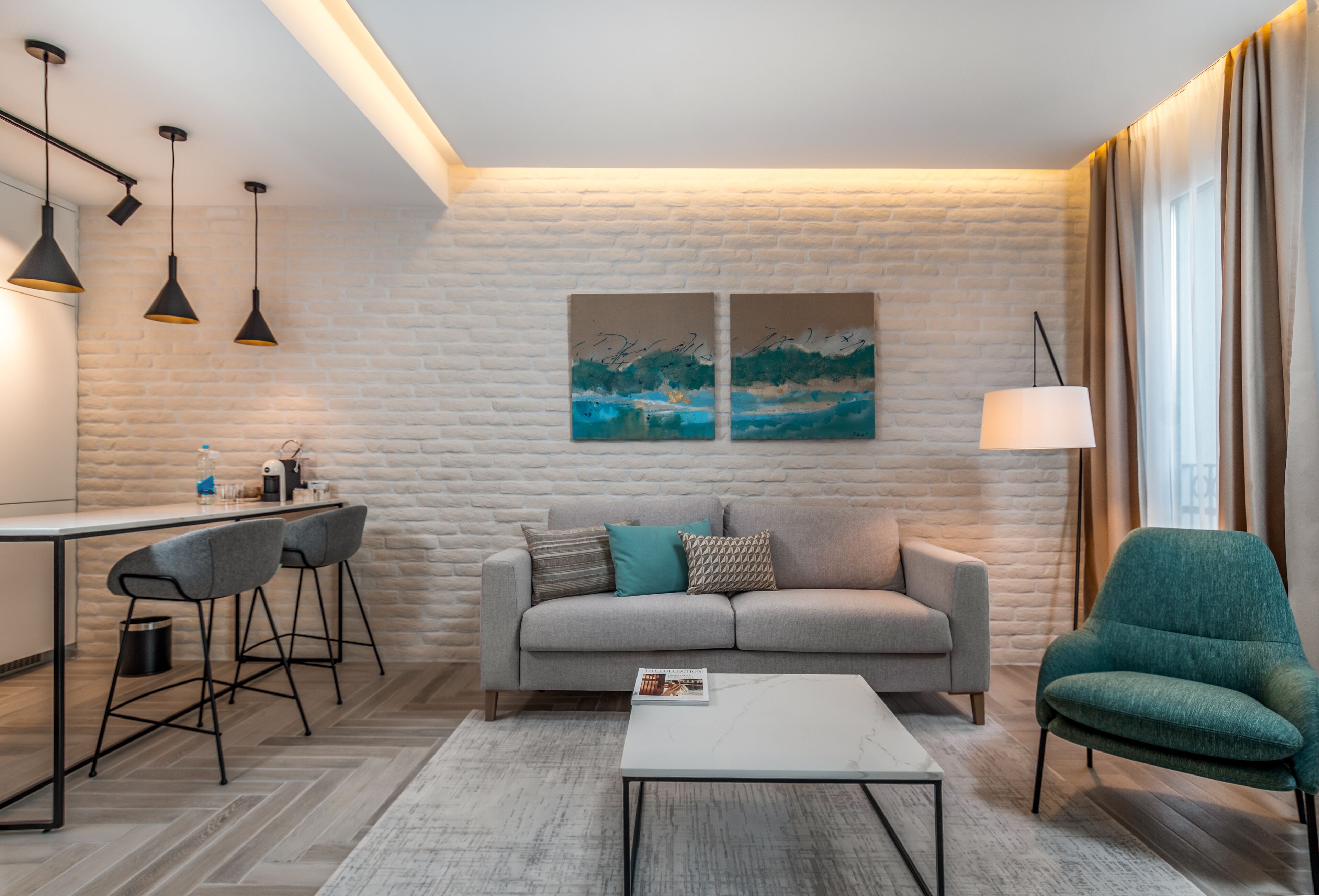 A modern living room with a sofa, a coffee table, and a dining area with bar stools. There are two paintings on the wall and a lamp on a side table.
