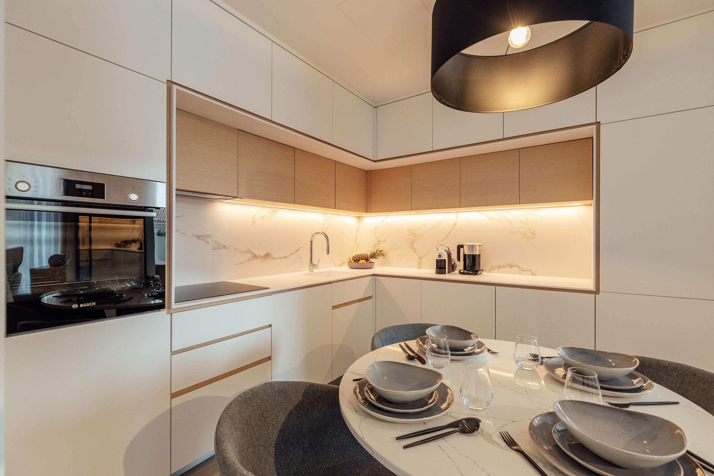 Una cocina moderna con horno Bosch, encimera de mármol y comedor con platos y cubiertos grises.