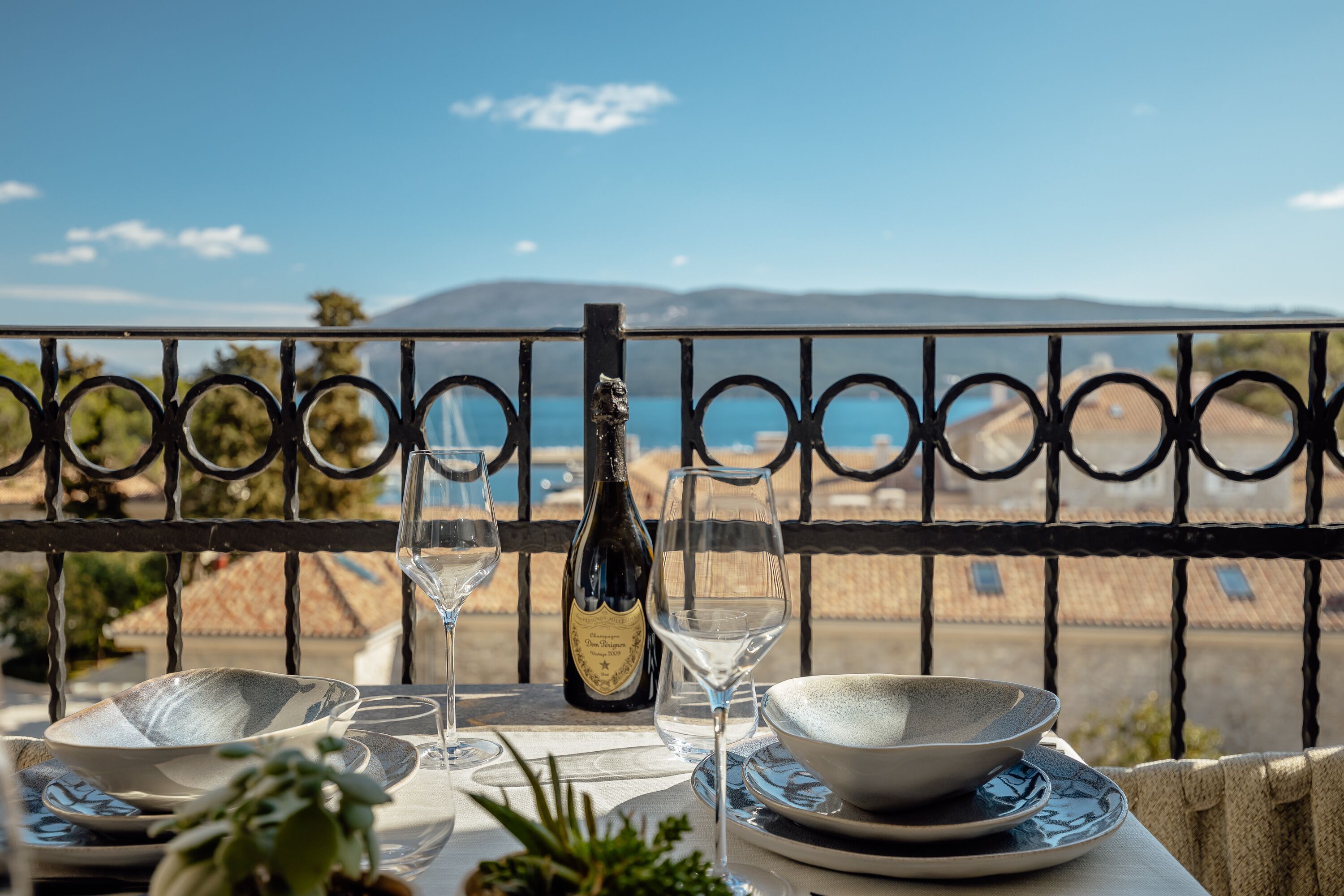 Une table dressée pour le dîner, avec une bouteille de vin, des verres et des bols, offrant une vue panoramique sur la mer et les montagnes.