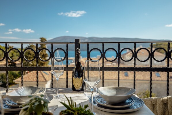 A table set for dining with a bottle of wine, glasses, and bowls, overlooking a scenic view of the sea and mountains.