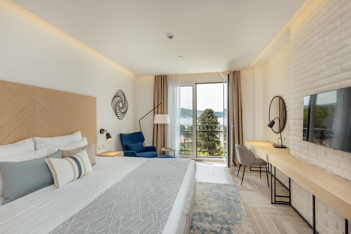 A modern hotel room with a large bed, a desk, a chair, and a balcony with a view.