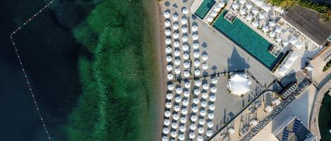 Vue aérienne d’un complexe hôtelier situé en bord de mer, avec une piscine, des parasols et des chaises longues.