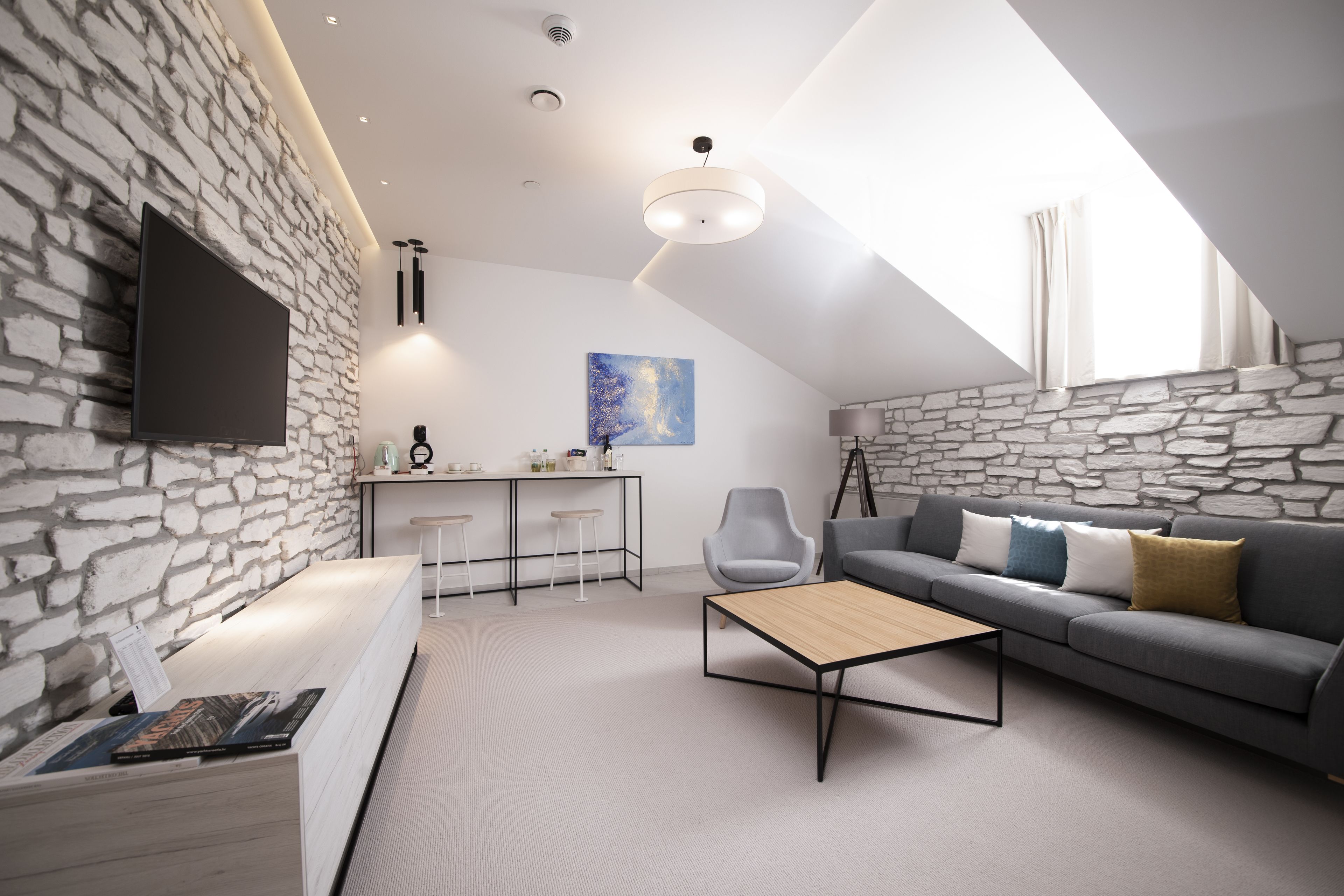 A modern living room with a stone wall, a flat-screen TV, a sofa with cushions, a coffee table, and a desk with stools.