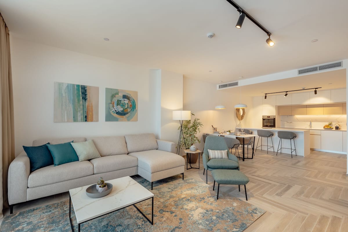 Ein modernes Wohnzimmer mit einer Couch, Sesseln, einem Couchtisch und einem Teppich. Es gibt einen Essbereich mit Tisch und Stühlen sowie eine Küche im Hintergrund.