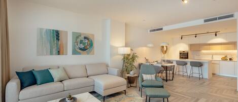 A modern living room with a sofa, armchairs, a coffee table, and a rug. There is a dining area with a table and chairs, and a kitchen in the background.
