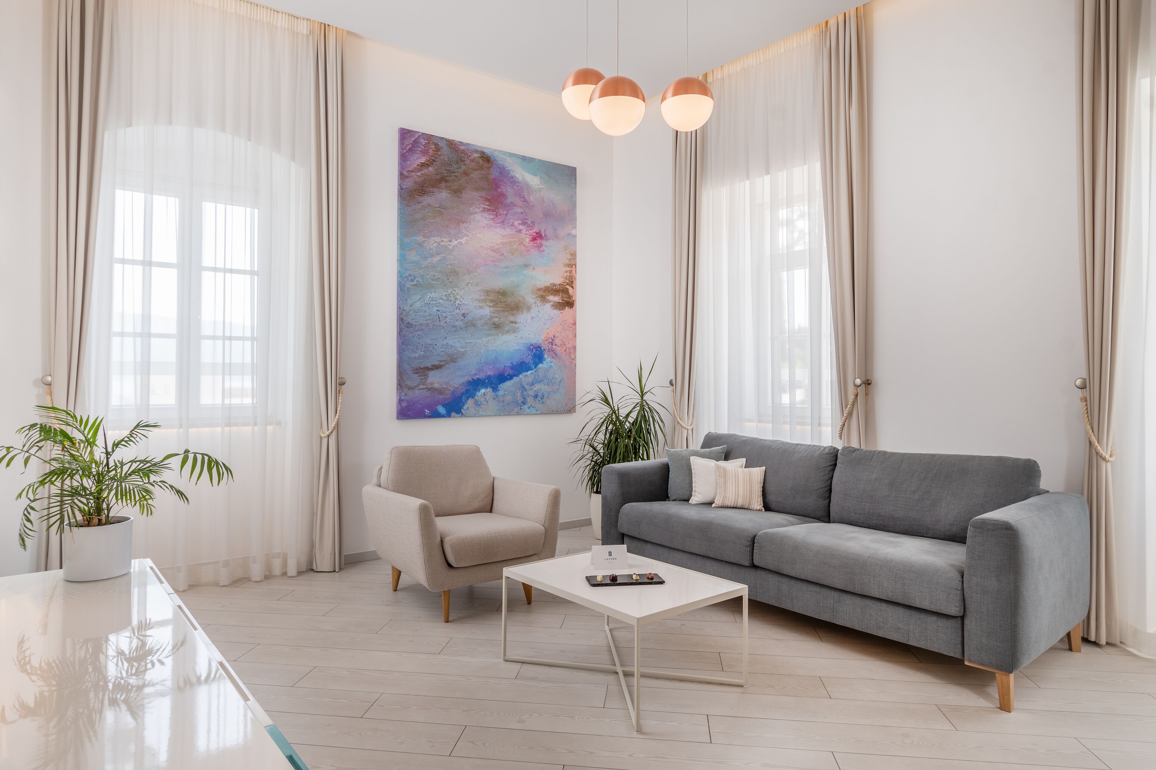 Een moderne woonkamer met een grijze bank, een witte salontafel, een beige fauteuil, en een groot abstract schilderij aan de muur.