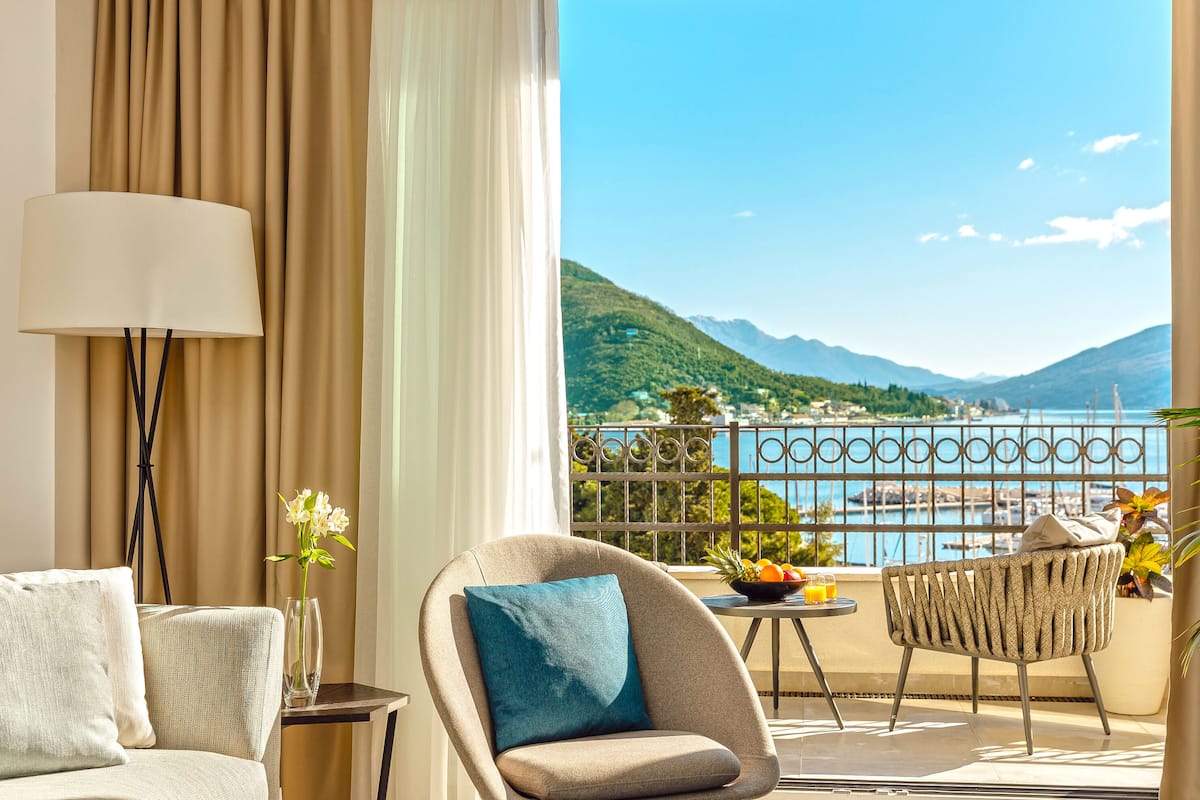 Ein Balkon mit Blick auf Berge und ein Gewässer, ein Sofa, ein Sessel, ein Tisch mit Früchten und eine Lampe.