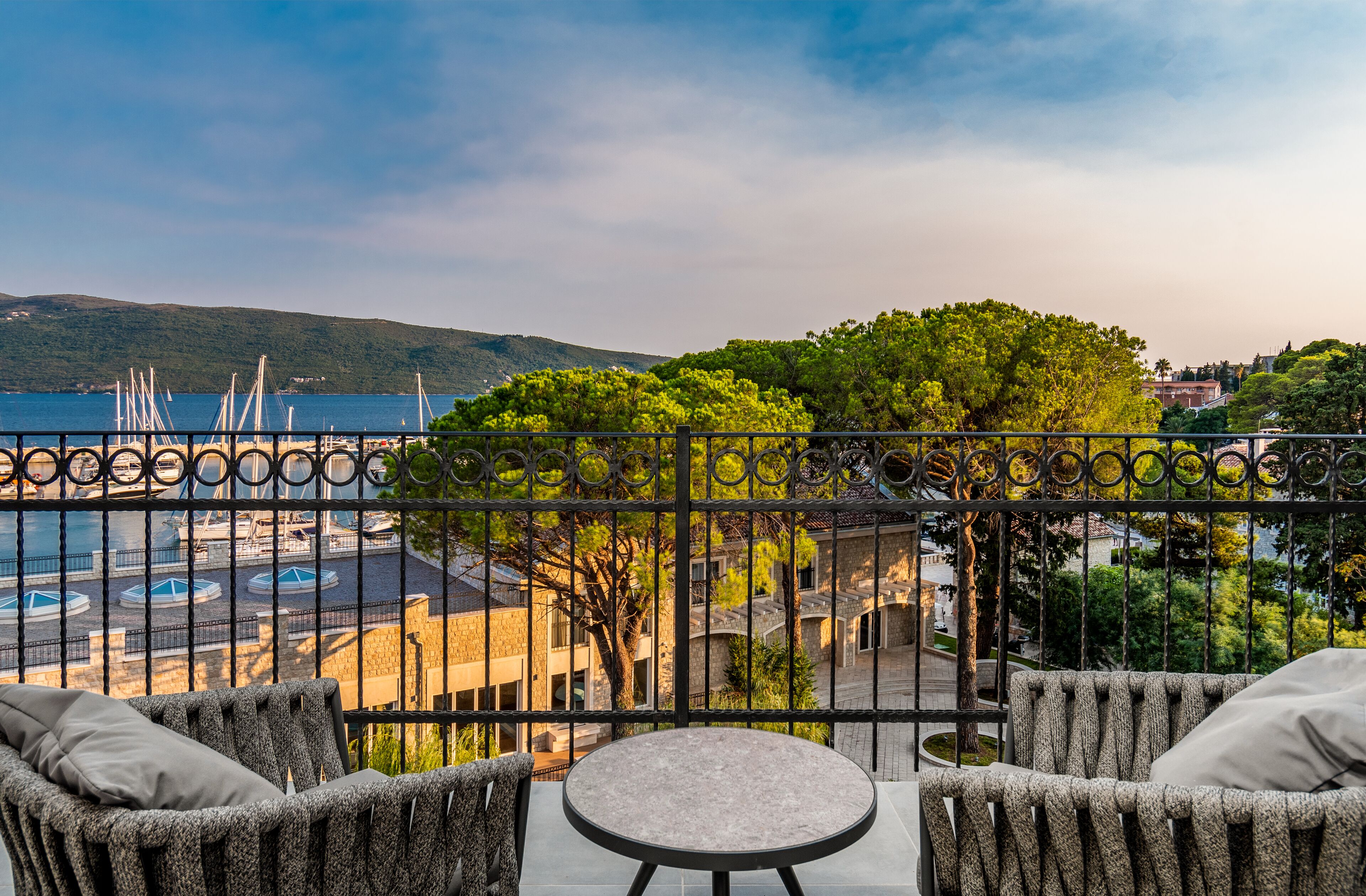 Un balcon agrémenté de meubles en osier, d’une table ronde et offrant une vue sur une marina avec des bateaux et un paysage montagneux.