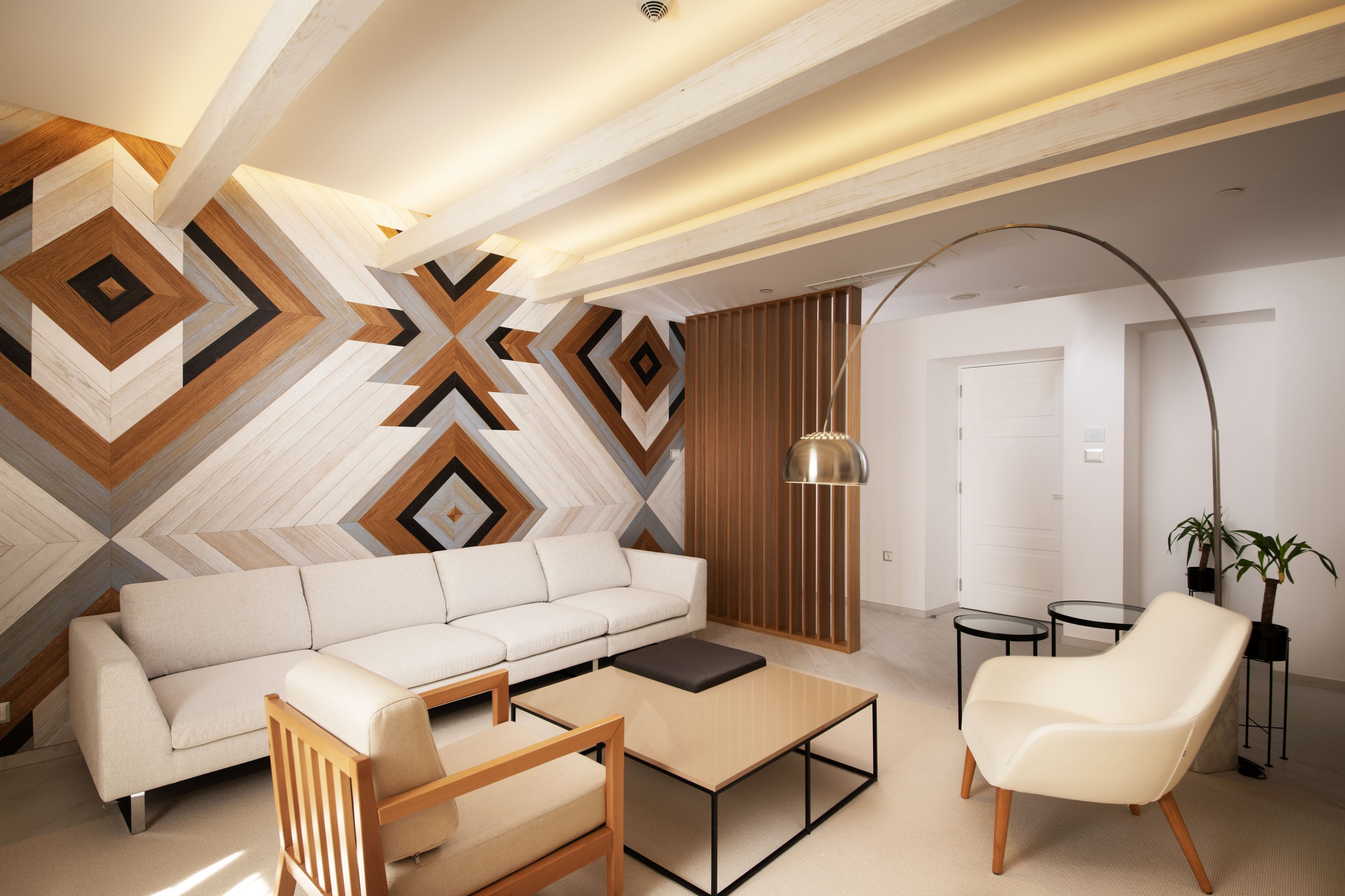 A modern living room with a white sofa, a wooden coffee table, a white armchair, and a geometric-patterned wall.