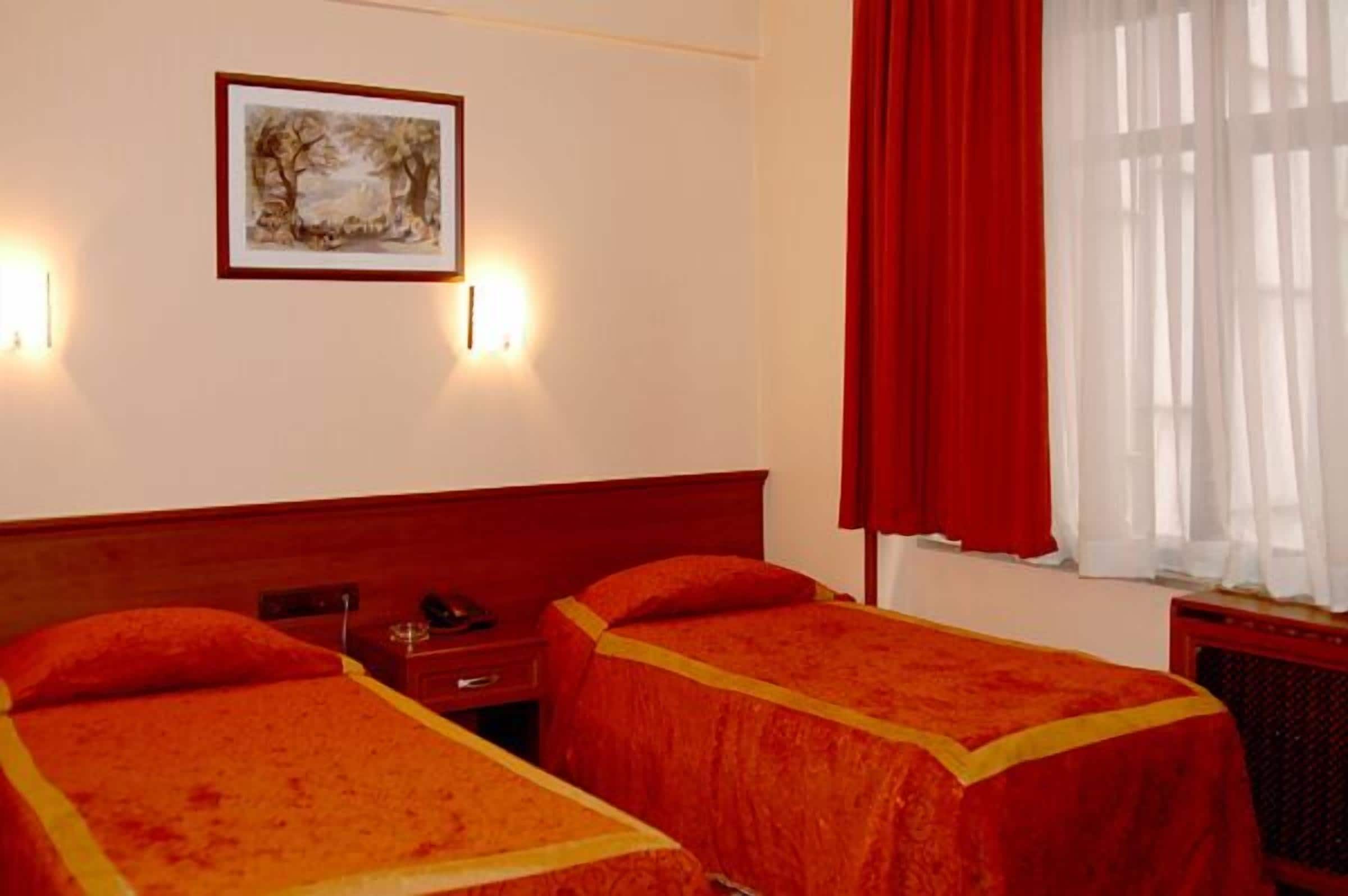 A hotel room with two single beds, a bedside table with a lamp, a framed picture on the wall, and a window with red curtains.