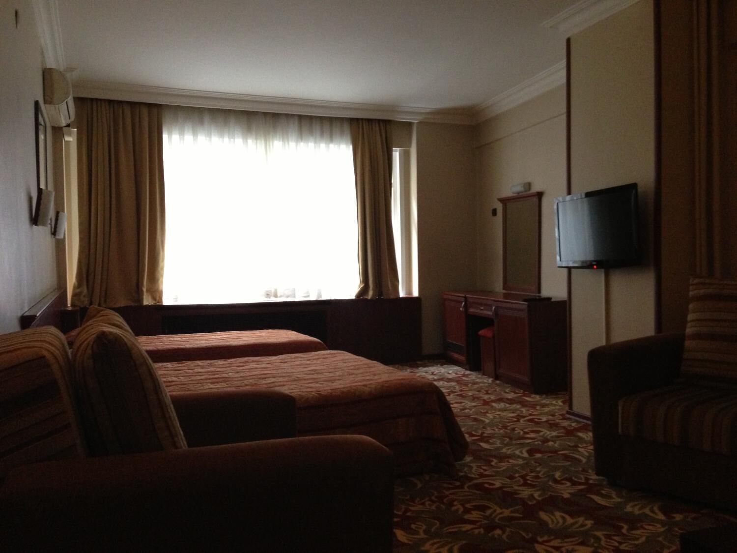 A hotel room with two beds, a desk, a chair, a television, and a large window with curtains.
