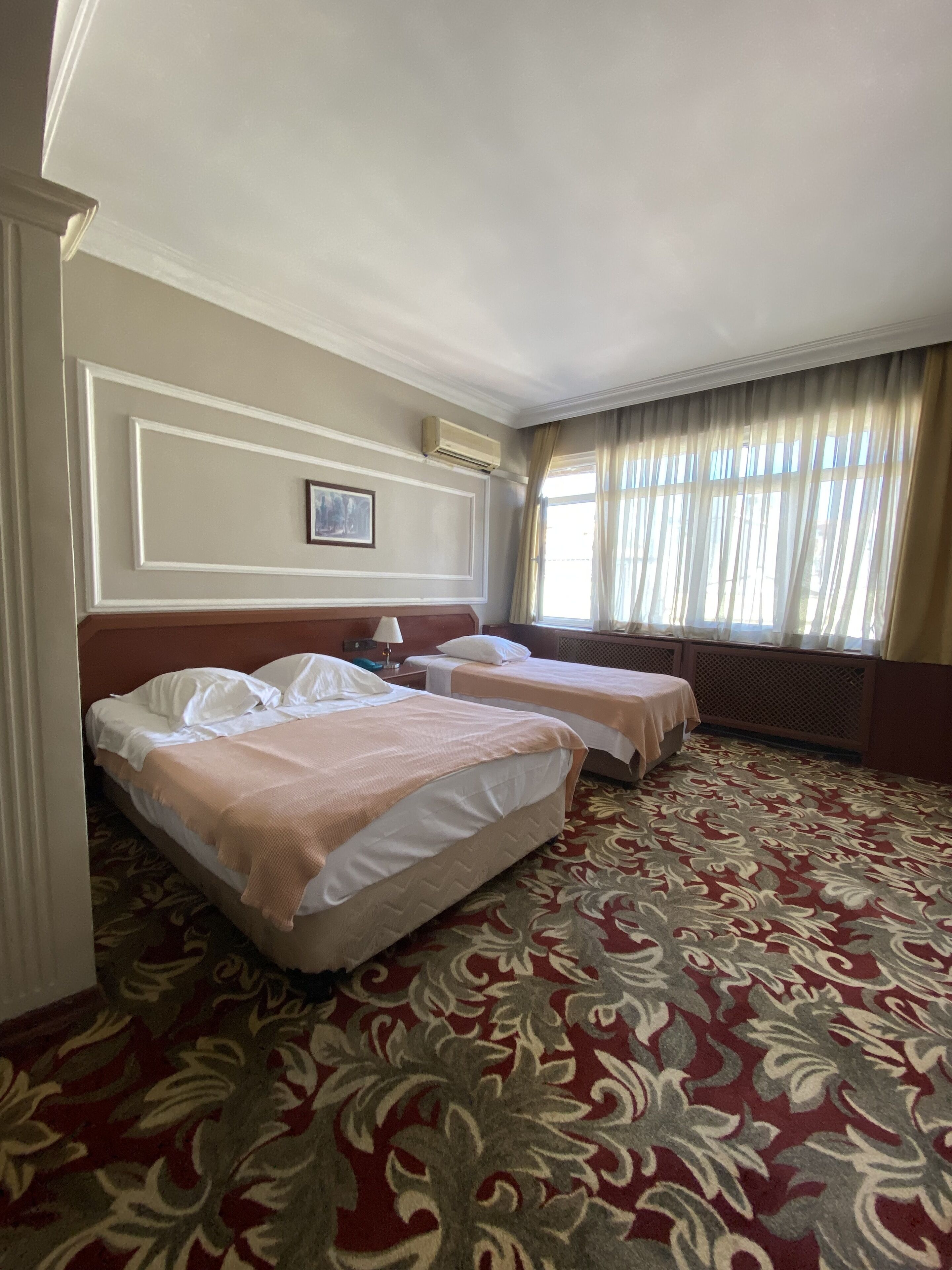 A hotel room with two beds, a patterned carpet, a window with curtains, and a painting on the wall.