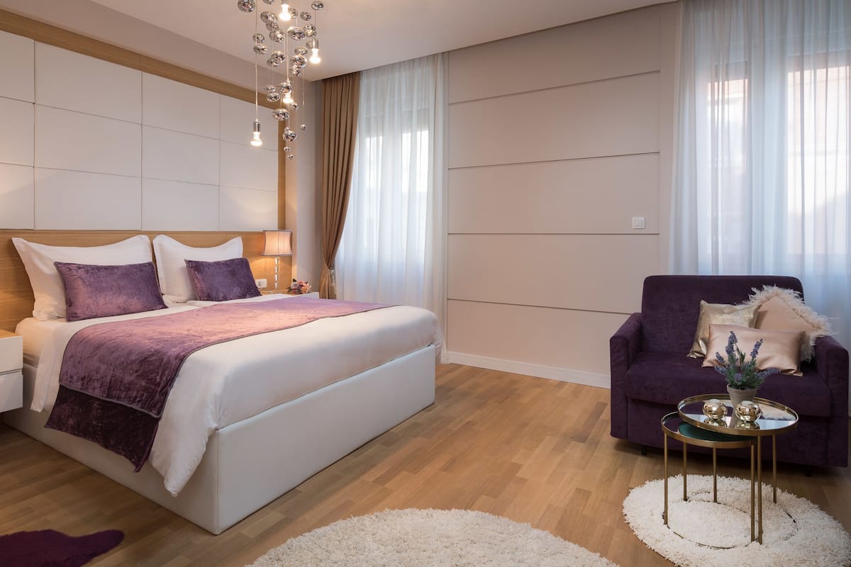 A modern bedroom with a large bed, bedside tables, a purple armchair, and a small round table with decorative items.