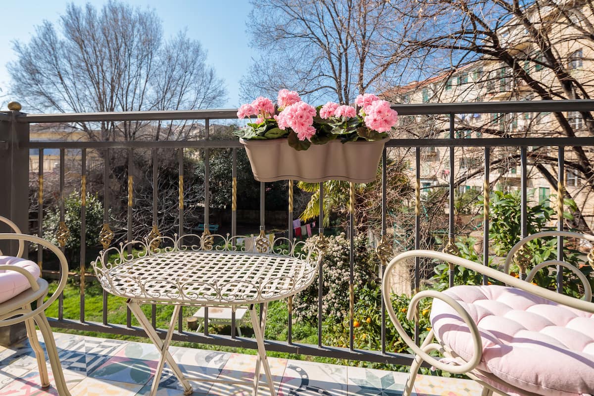 A balcony with a metal table and chairs, a hanging flower pot with pink flowers, and a view of a garden and buildings in the background.