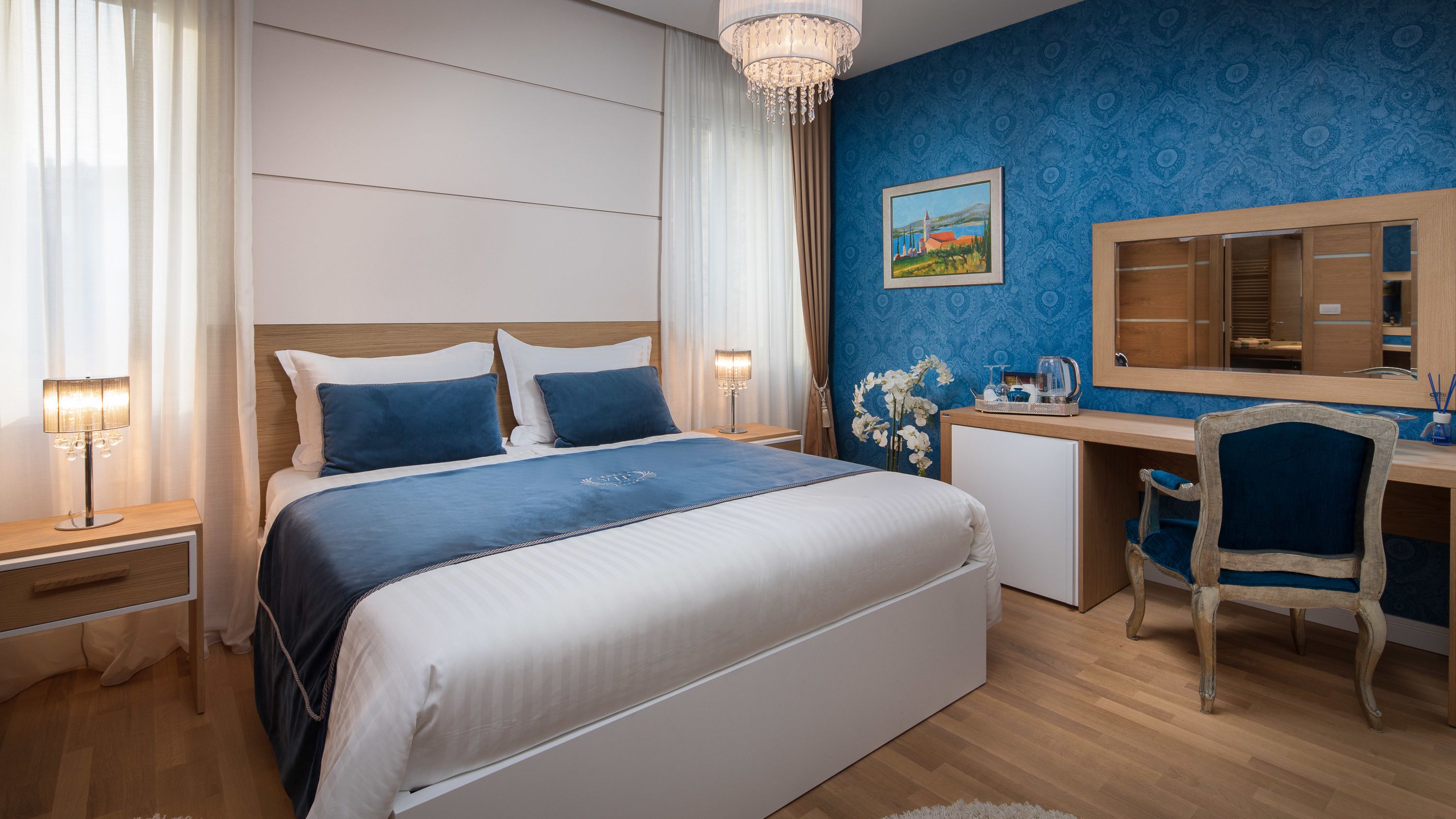 Ein Hotelzimmer mit einem großen Bett, einem Schreibtisch, einem Sessel, einem kleinen Kühlschrank und einer Kronleuchter.