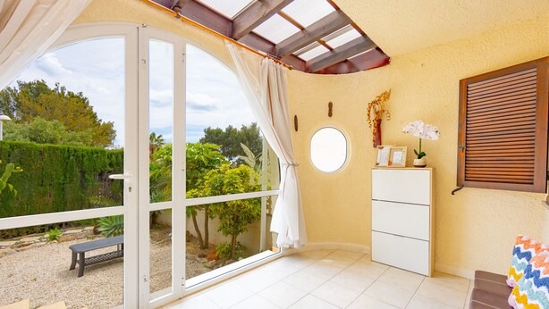 Una habitación con puerta de vidrio corrediza que da a un jardín, un pequeño aparador blanco, un banco y una manta colorida sobre un sofá marrón.