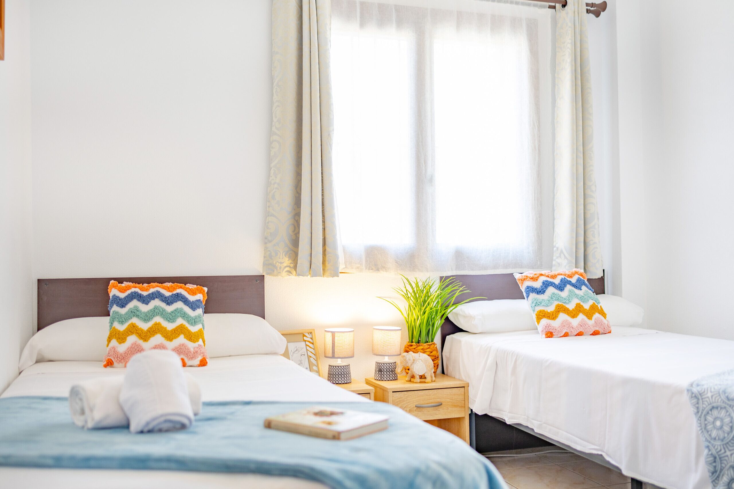 Habitación de hotel con dos camas, cada una con un cojín colorido en zigzag, una mesita de noche pequeña con una lámpara, una decoración de piña y una ventana con cortinas traslúcidas.