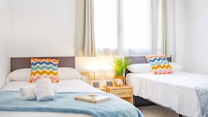 Habitación de hotel con dos camas, cada una con un cojín colorido en zigzag, una mesita de noche pequeña con una lámpara, una decoración de piña y una ventana con cortinas traslúcidas.