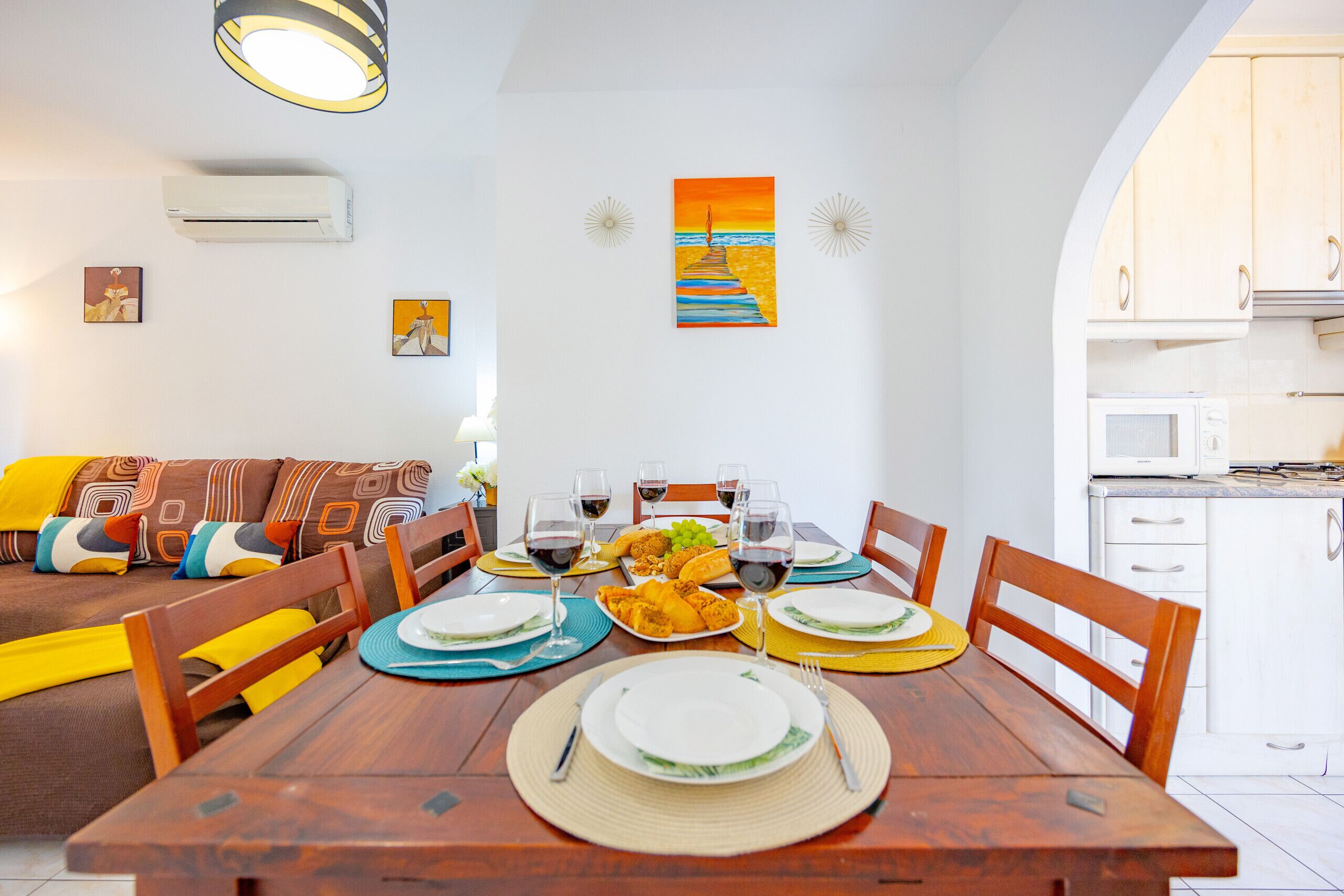 A dining area with a wooden table set for four, surrounded by chairs. There is a sofa with colorful cushions in the background.