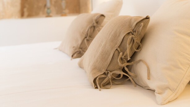 A bedroom with a stone wall, wooden beams, and a bed with beige pillows.