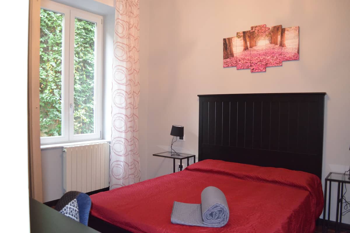 A bedroom with a red bedspread, a rolled towel on the bed, a black headboard, a window with a view of trees, a wall-mounted picture, and a small table with a lamp.