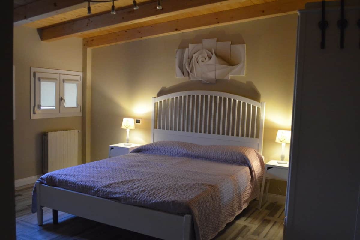 A bedroom with a wooden ceiling, a bed with a white headboard, two bedside lamps, and a large abstract wall decoration.