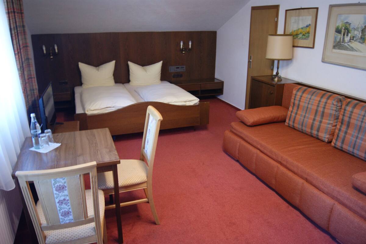 A hotel room with a double bed, a sofa, a wooden table with a chair, a desk, and a lamp.