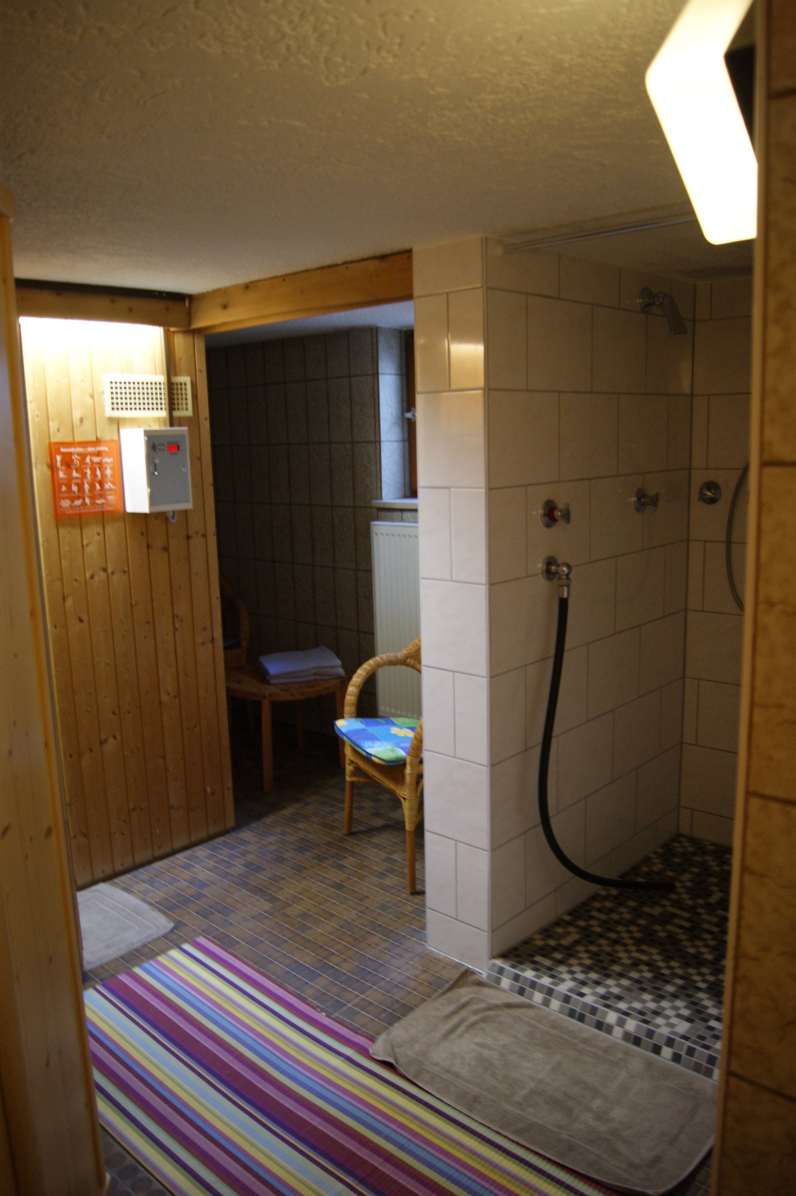 A small, compact bathroom with a shower, a wooden chair, and a striped rug.