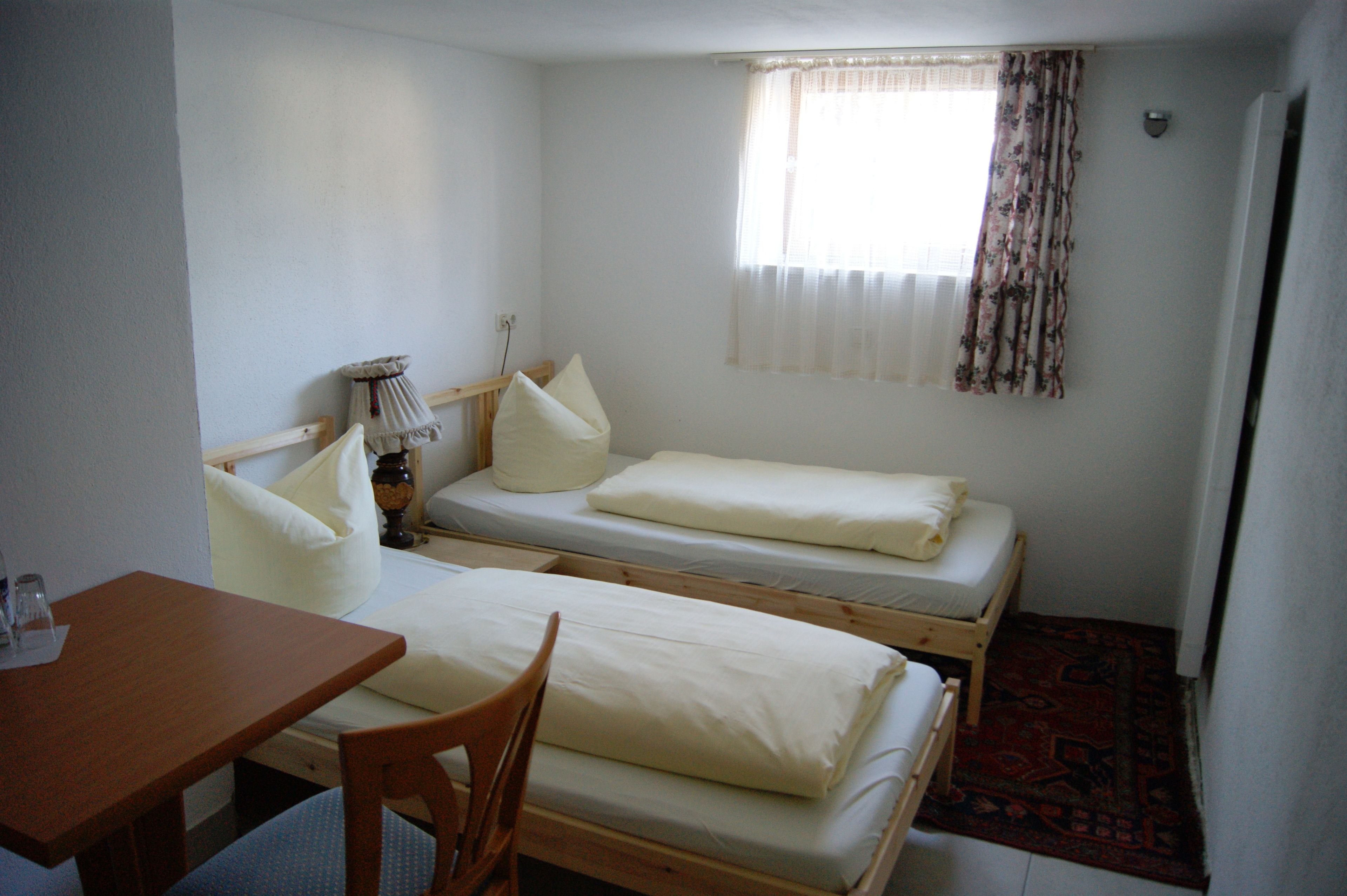 A small, simple room with two single beds, a wooden table, and a chair.