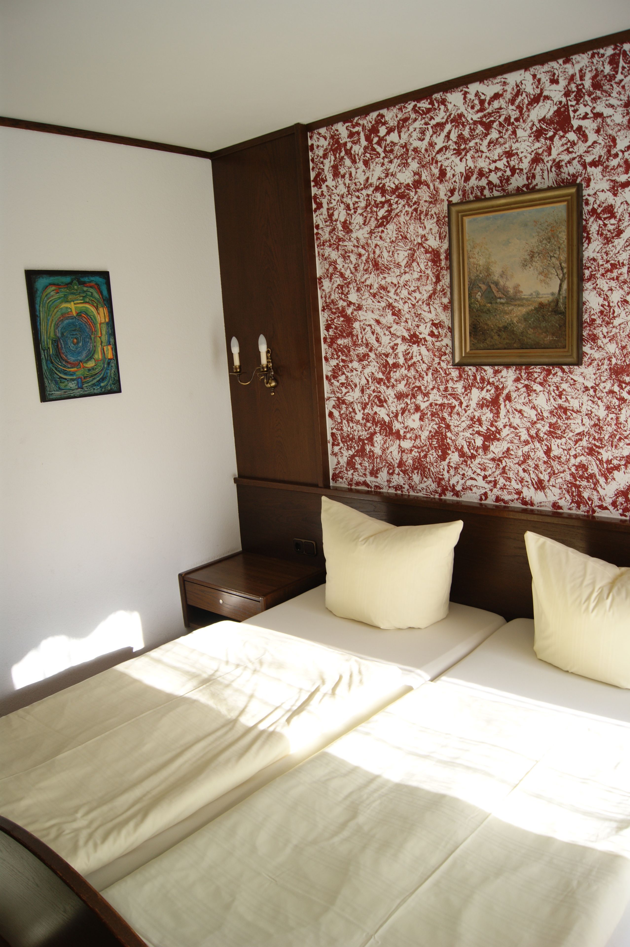 A bedroom with a bed, a nightstand, a wooden wardrobe, a painting, and a colorful abstract artwork on the wall.