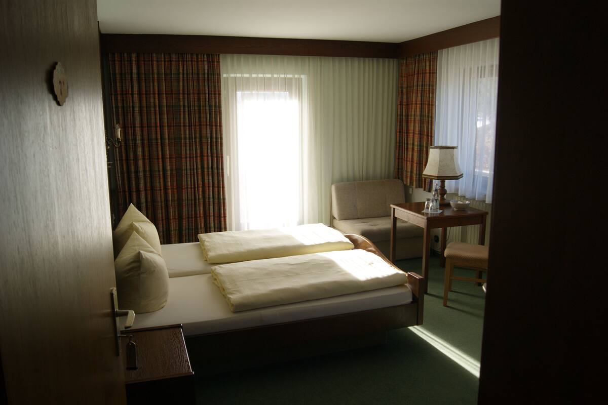 A hotel room with a bed, a chair, a small table with a lamp, and a window with curtains.