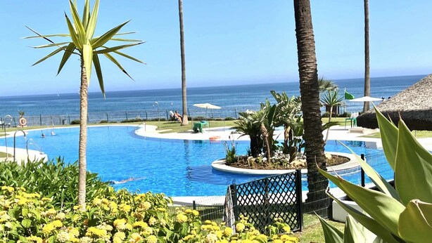 A pool area with palm trees, a view of the ocean, and a thatched-roof structure. - MI CAPRICHO 16 BEACHFRONT APARTMENT (Mijas)