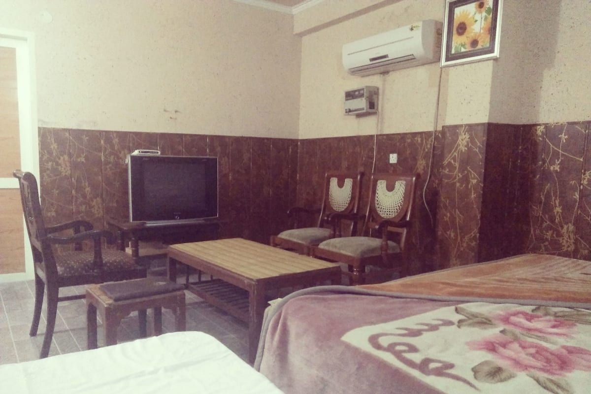 A hotel room with a bed, a television, two chairs, a small table, and an air conditioning unit.
