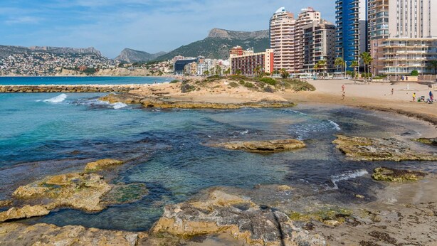 A coastal cityscape with a rocky beach, clear blue sea, and high-rise buildings in the background. - Apartament Arenal 1 - PlusHolidays (Calpe)