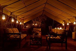 Un espace aménagé sous une tente, avec un canapé, des fauteuils et une table, le tout placé sous une sorte de dais décoré de guirlandes lumineuses.