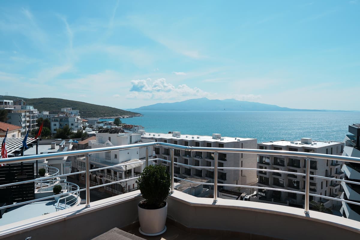A balcony with a potted plant, overlooking a coastal cityscape with buildings and a clear blue sky.