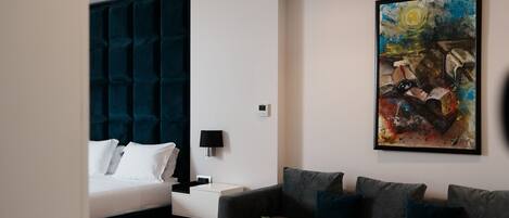 A modern hotel room with a bed, a sofa, a coffee table, a painting, and a ceiling vent.