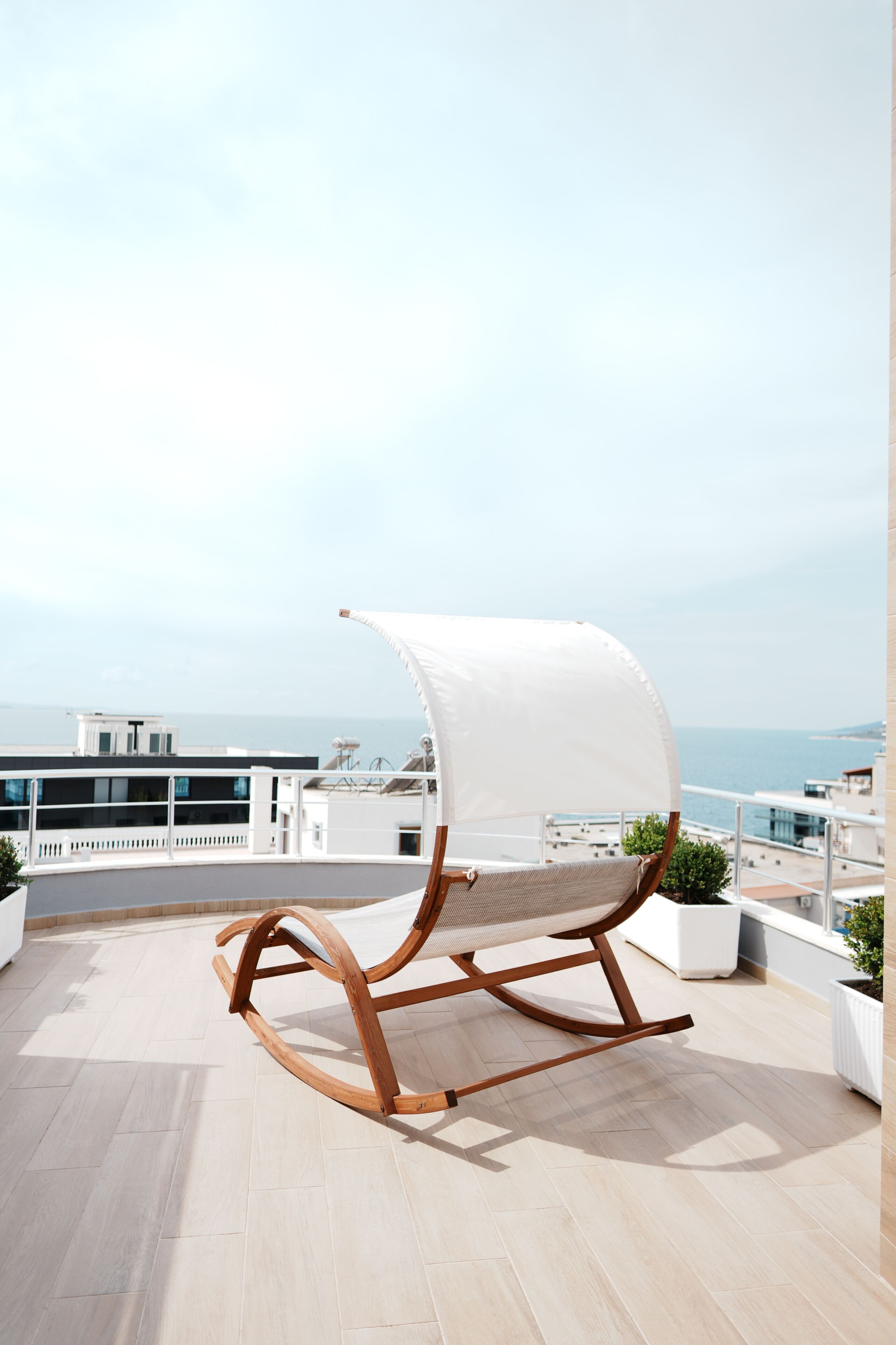 A wooden rocking chair with a white canopy on a balcony with a view of the sea and buildings.