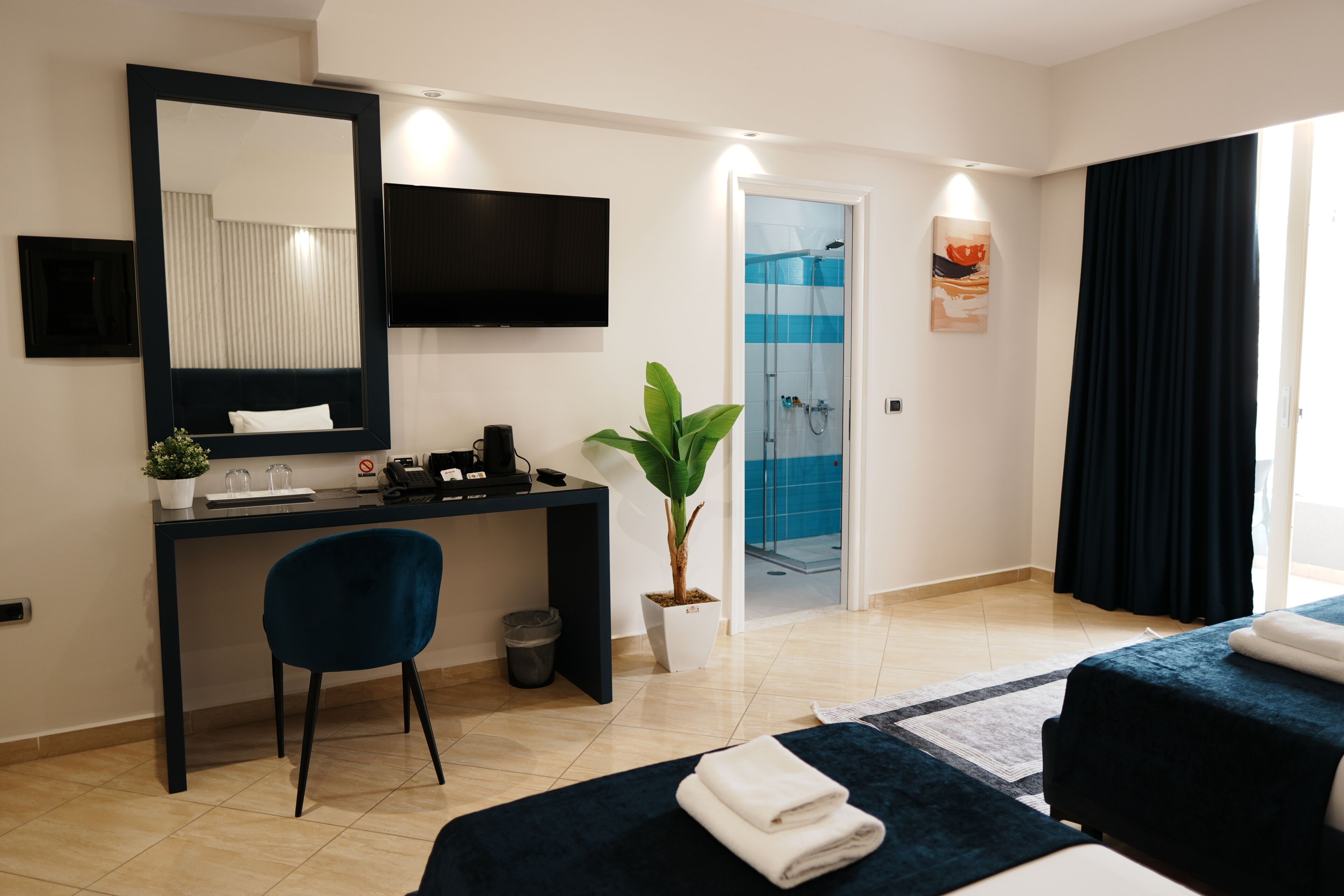 A hotel room with a desk, chair, two beds, a TV, a plant, a shower, and a window with curtains.
