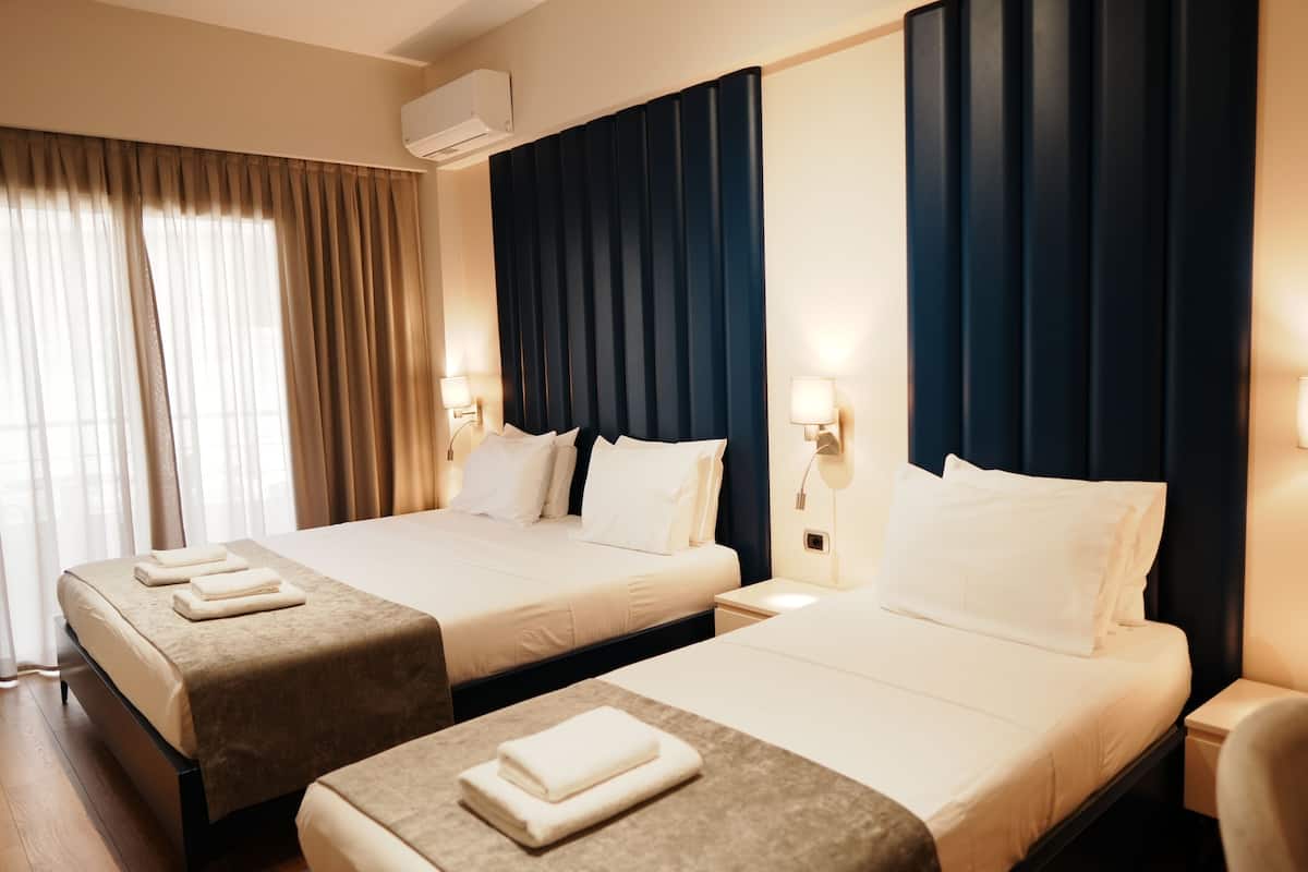 A hotel room with two beds, a large window with curtains, a wall-mounted air conditioner, and bedside lamps.