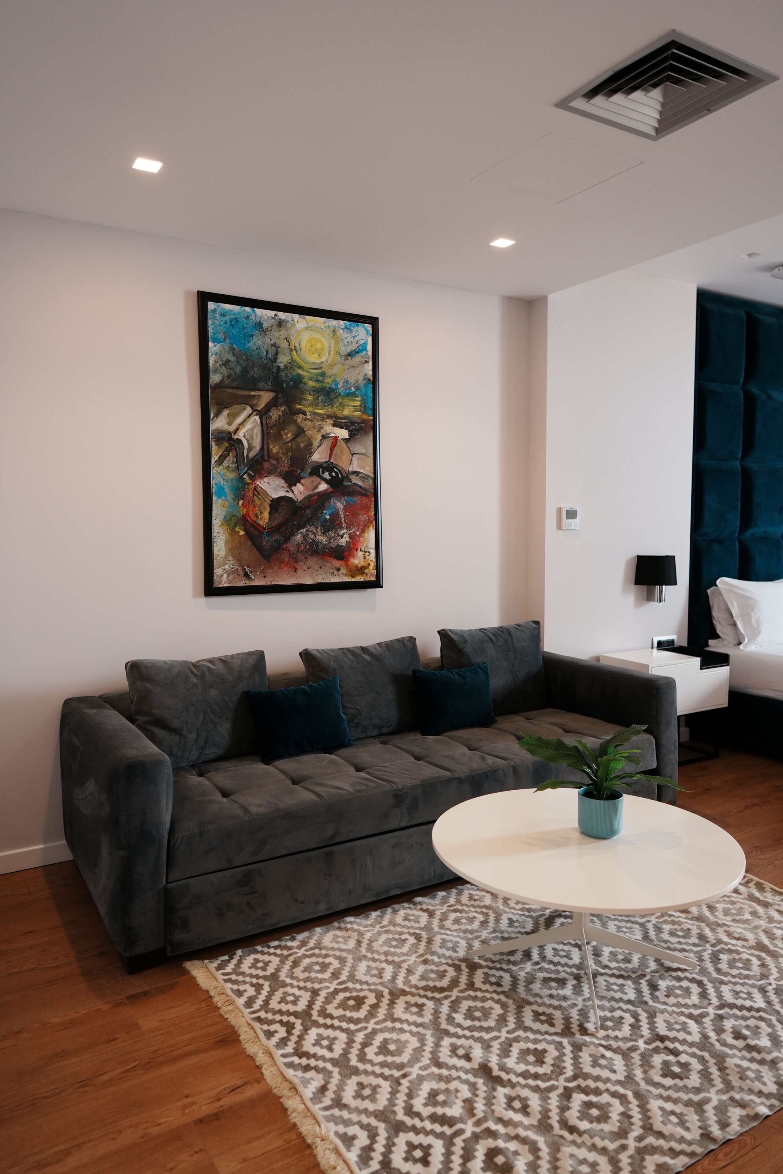 A modern living room with a grey sofa, a round coffee table, a patterned rug, a painting on the wall, and a blue curtain.