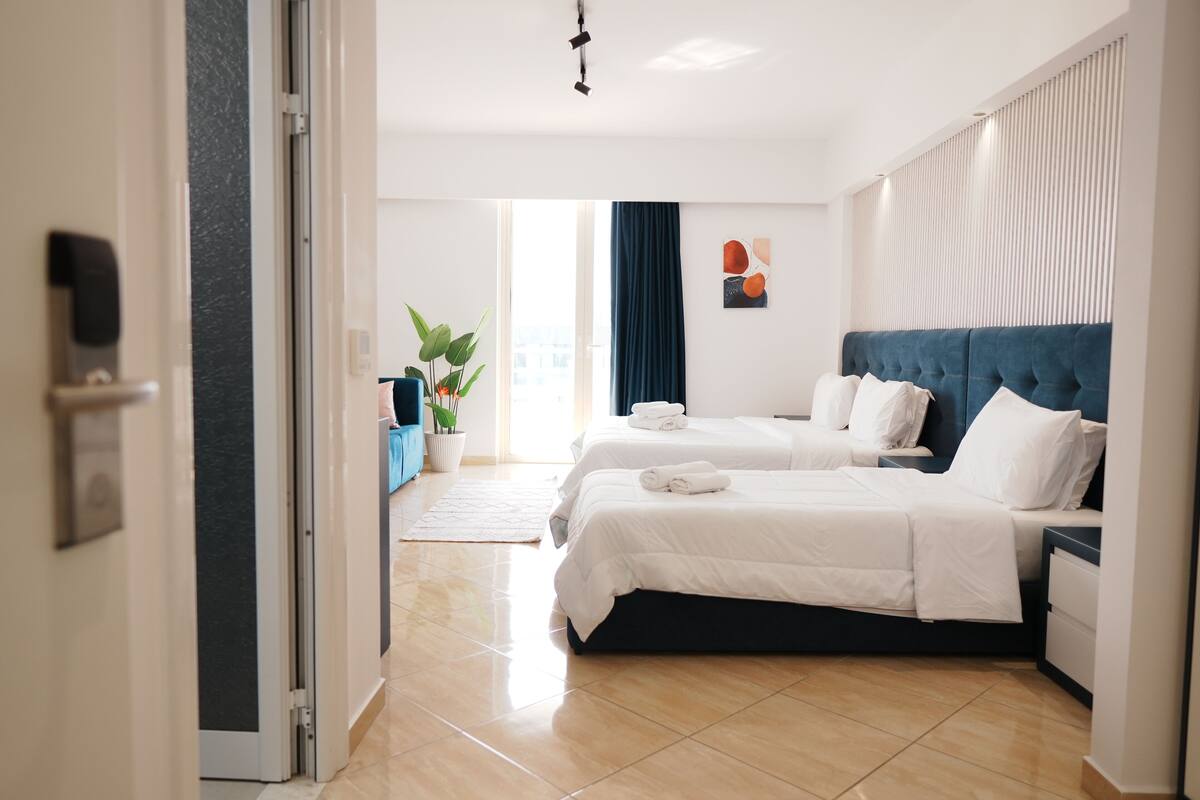 A hotel room with two beds, a potted plant, a blue sofa, and a view of the city.