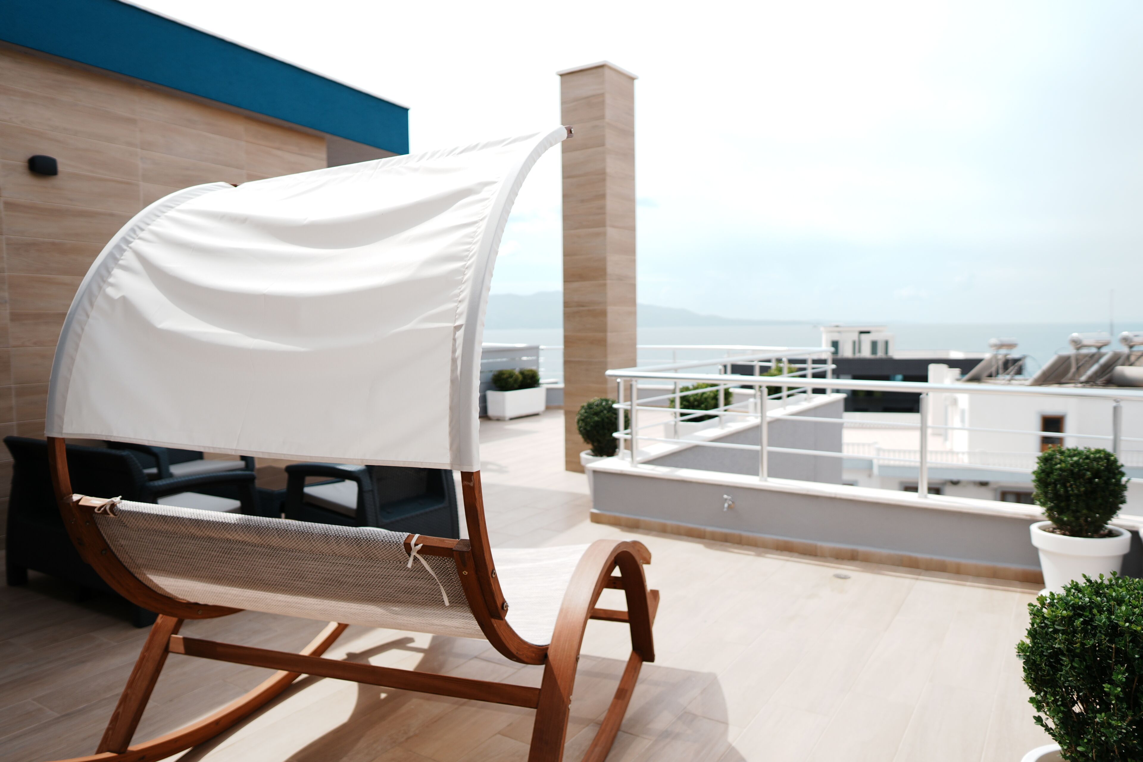 Un fauteuil à bascule en bois, surmonté d’un auvent blanc, se trouve sur une terrasse en toit, d’où l’on peut admirer la mer.