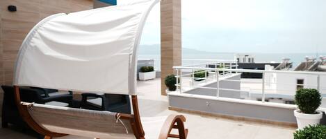 Un fauteuil à bascule en bois, surmonté d’un auvent blanc, se trouve sur une terrasse en toit, d’où l’on peut admirer la mer.