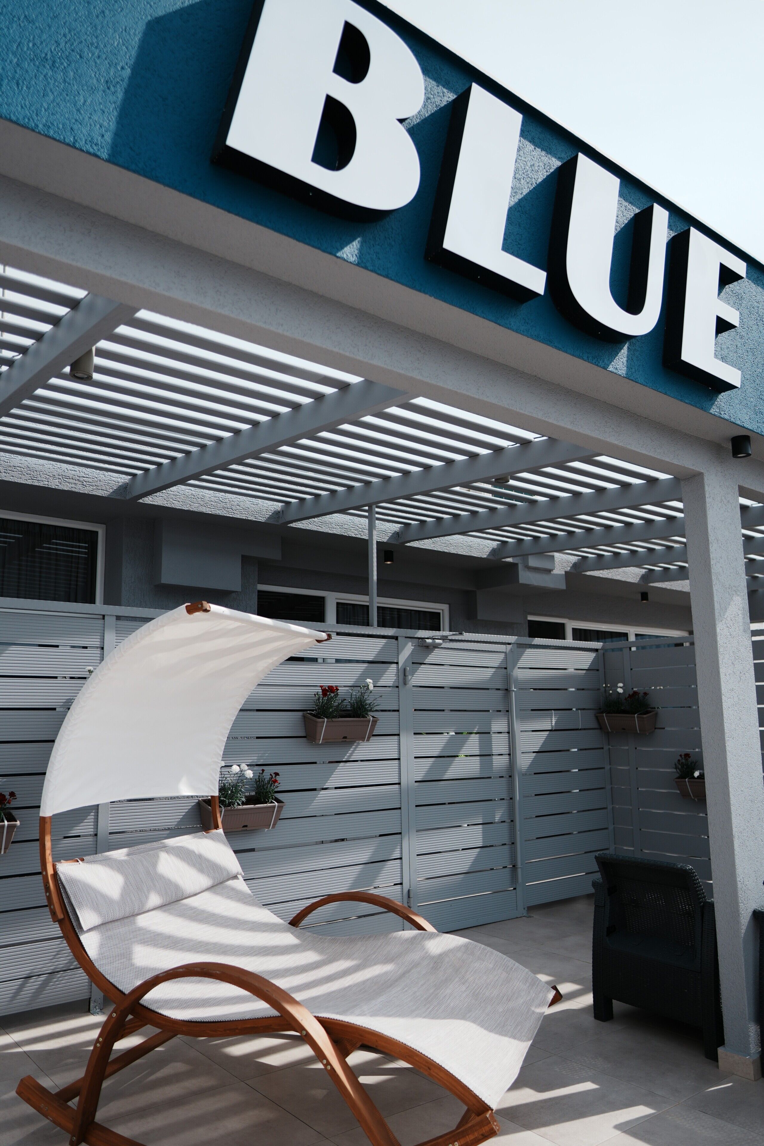 Un espacio de estar al aire libre con techo, con una mecedora y una mesita, una pared azul con la palabra 'BLUE' en letras grandes, y una cerca blanca con plantas en macetas.