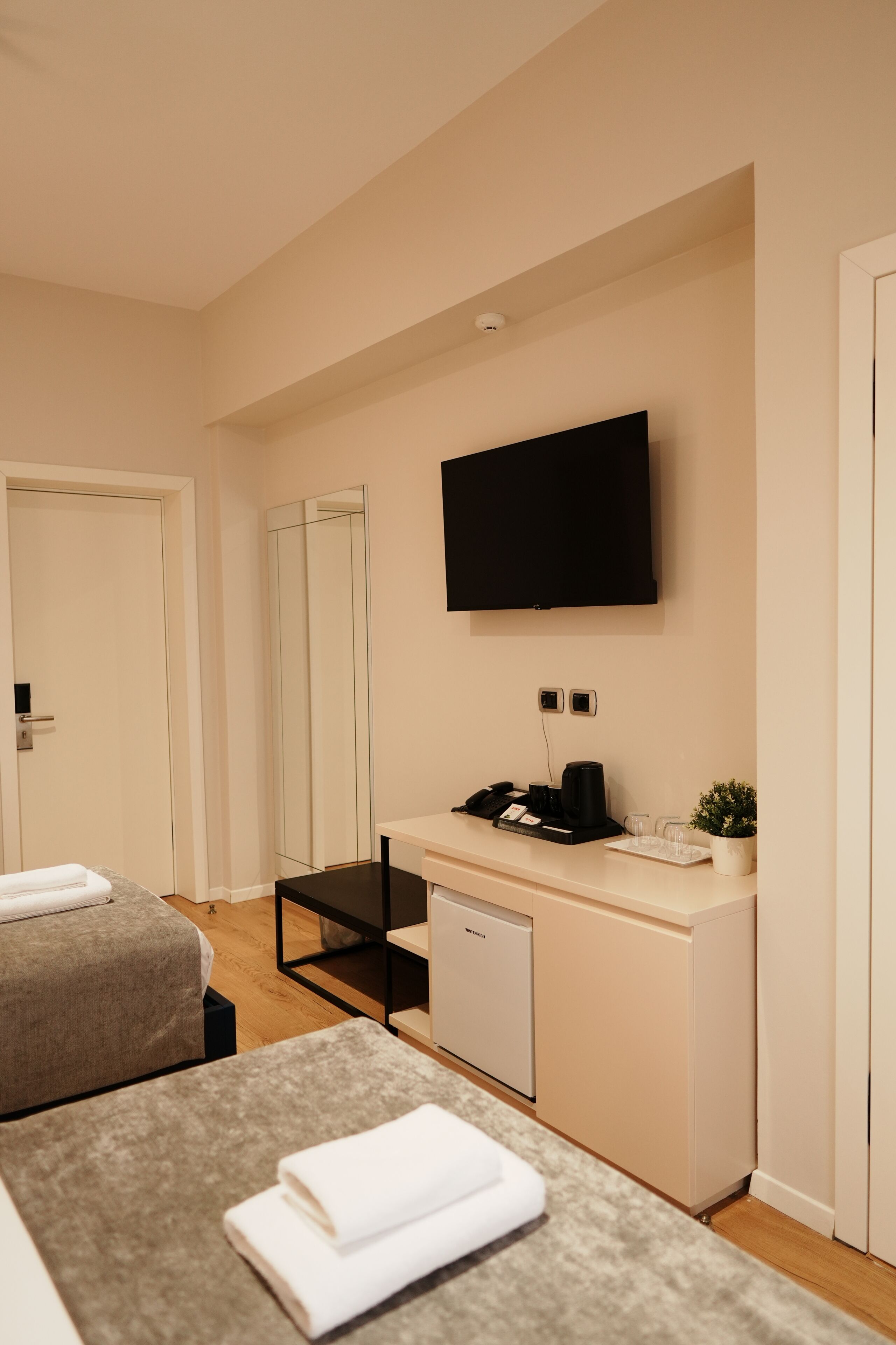 A hotel room with a bed, a desk with a flat-screen TV, a small kitchenette, and a mirror.