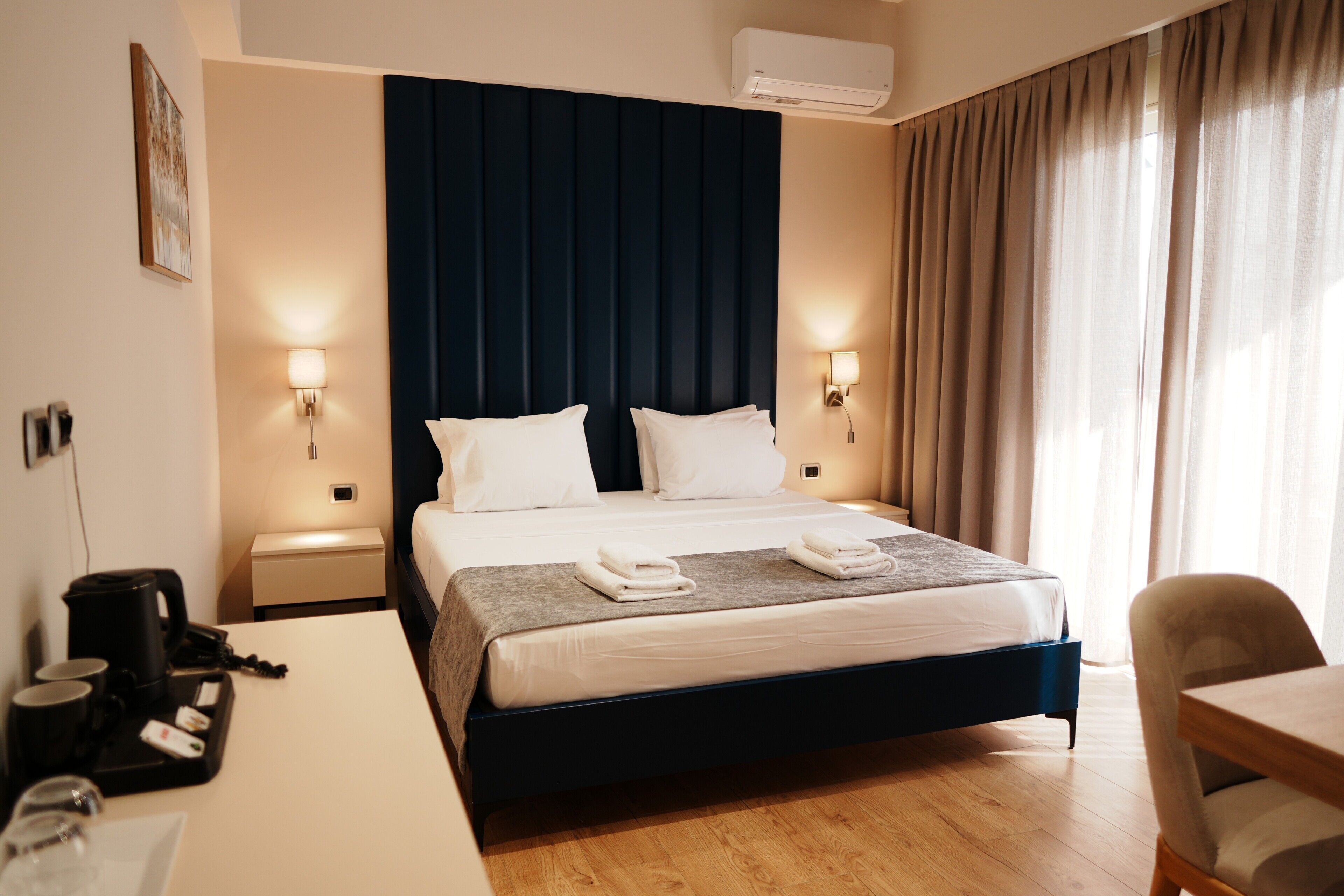 Quarto de hotel com cama, mesas de cabeceira, escrivaninha, cadeira e janela com cortinas.