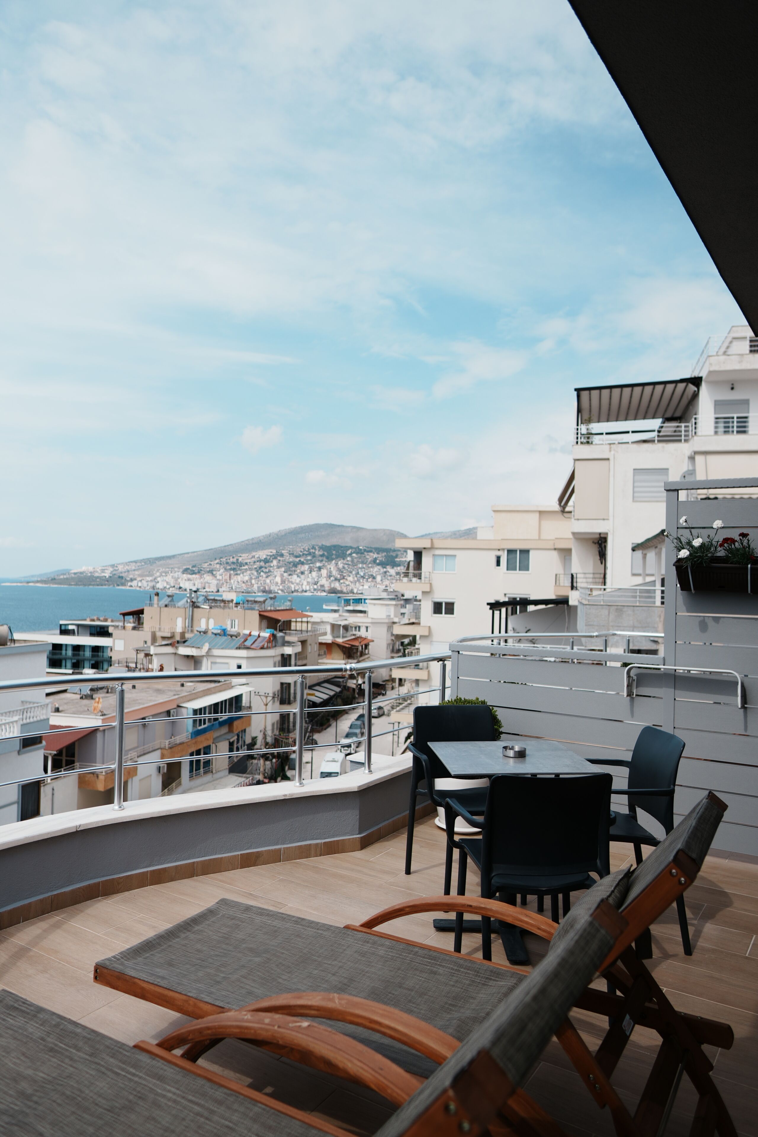 A balcony with a view of the sea and buildings, a table with chairs, and a wooden chair.