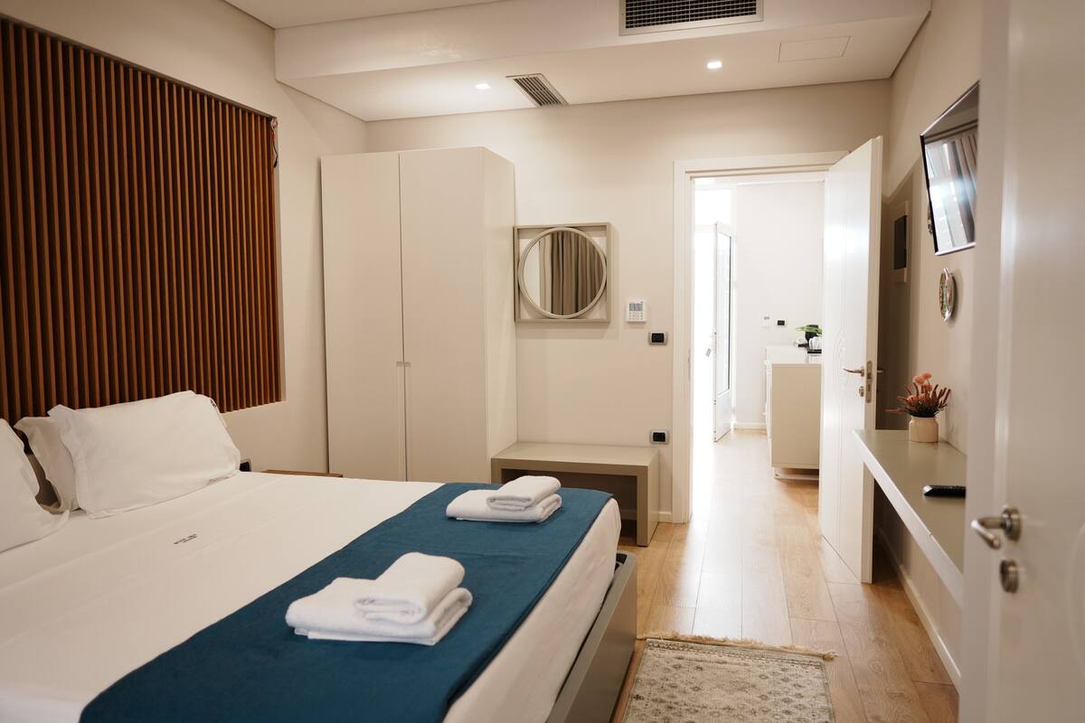 A modern hotel room with a bed, bedside tables, a mirror, a wardrobe, and a bathroom visible in the background.