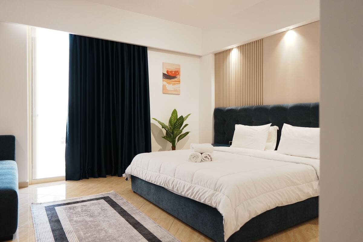 A modern hotel room with a large bed, a dark blue headboard, a potted plant, and a view of the outdoors.